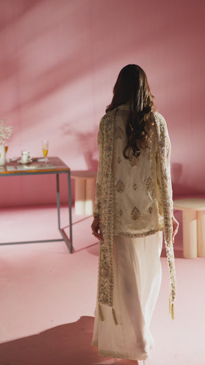 Video in which camera begins with soft focus on pink backdrop, then shifts to model turning gracefully over her shoulder in Reyna crème angrakha, gold and pastel embroidery catching the light. 
