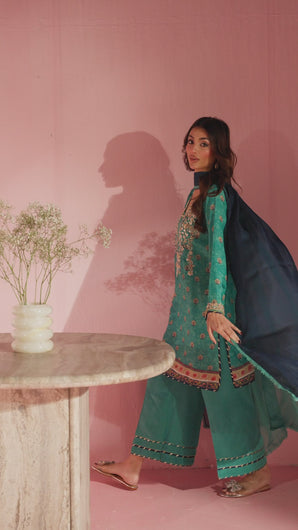 A video in which camera pans across soft pink backdrop as model turns candidly to the side, turquoise kameez embroidered with gold and pink floral motifs catching the light, wide‑leg trousers visible at hem. 
